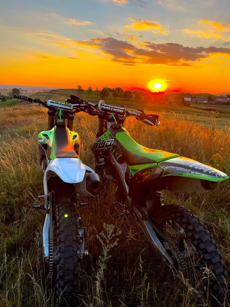 Rear view of two green dirt bikes side by side in a grassy field at sunset.