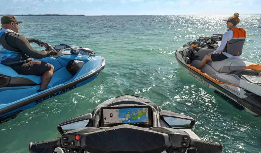 POV from a personal watercraft showing its dashboard map as two other jet skis with life-jacketed riders float nearby on calm, green-blue sea under a bright horizon.