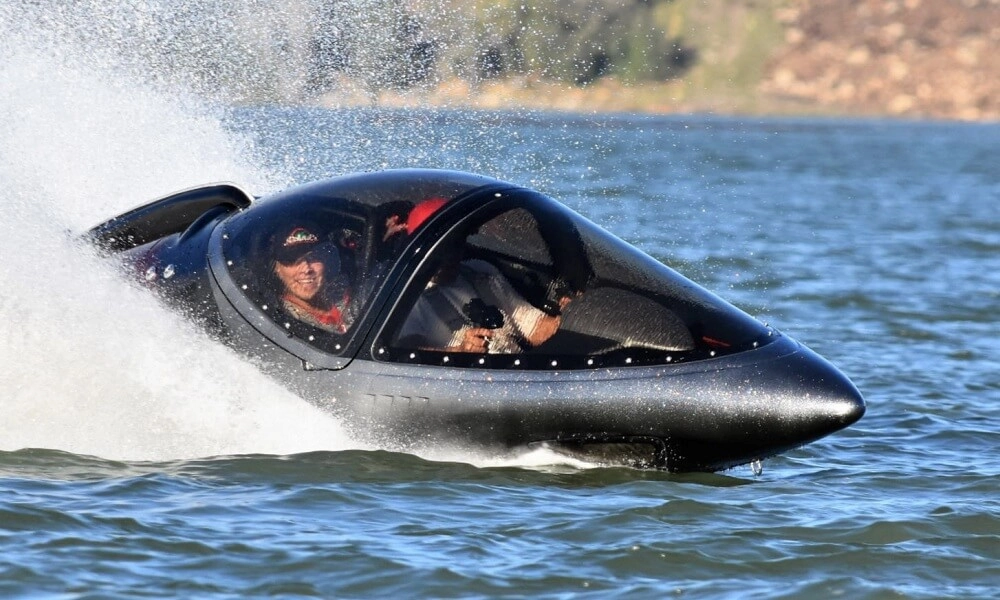 Black Seabreacher Z watercraft speeds across a lake; two riders sit under a clear bubble canopy as spray flies.